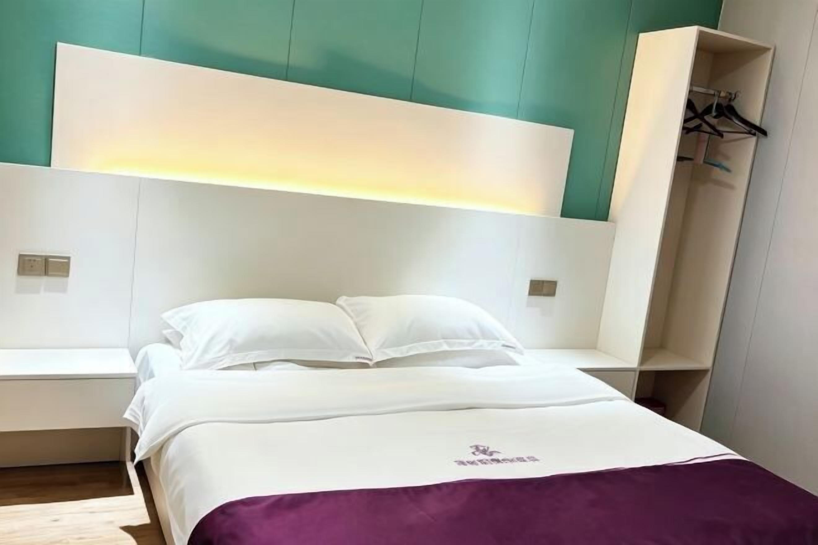 Un lit avec une literie blanche et un plaid violet, une table de chevet et une armoire avec un miroir.