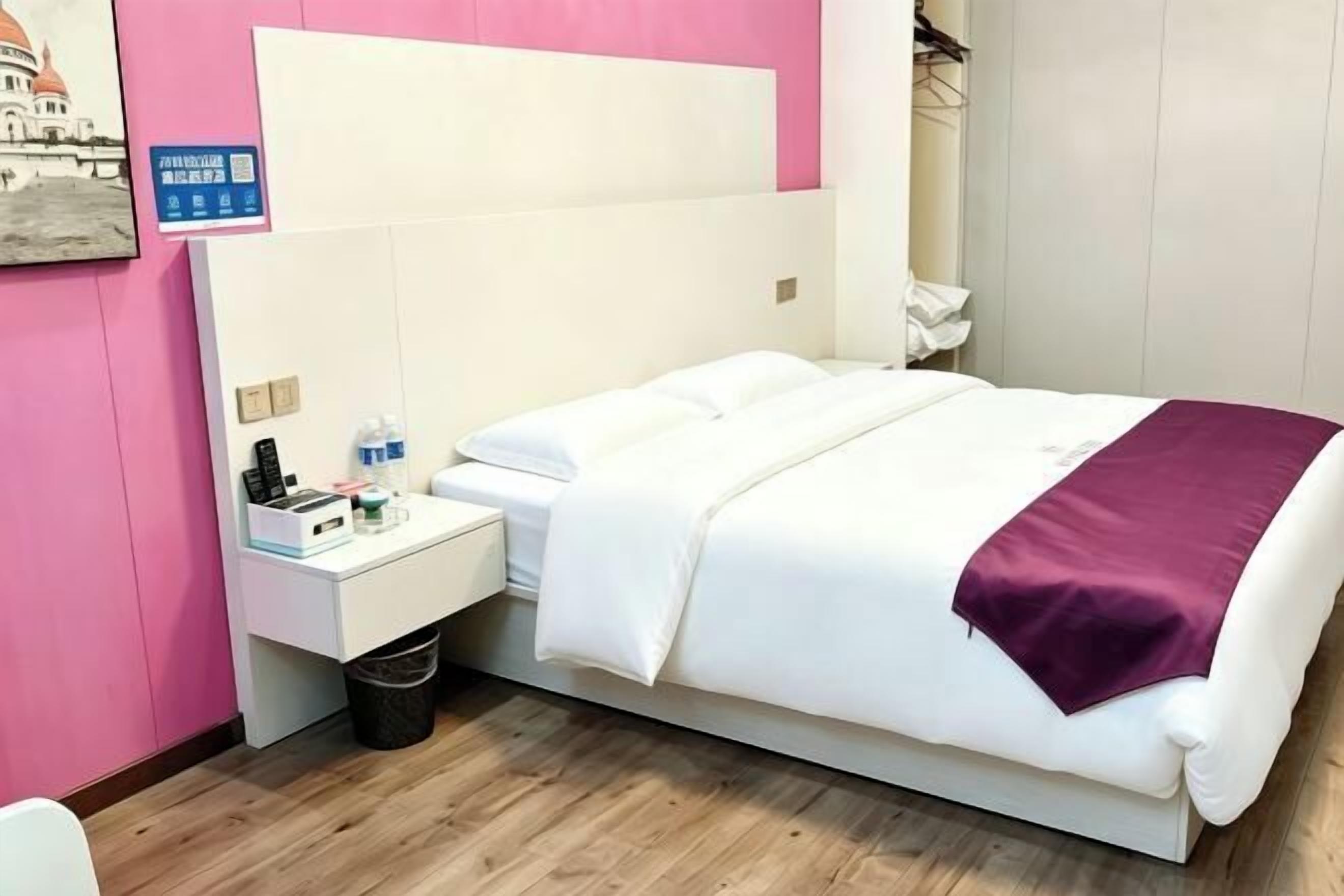 A hotel room with a bed, a nightstand, a trash bin, and a wall-mounted shelf.