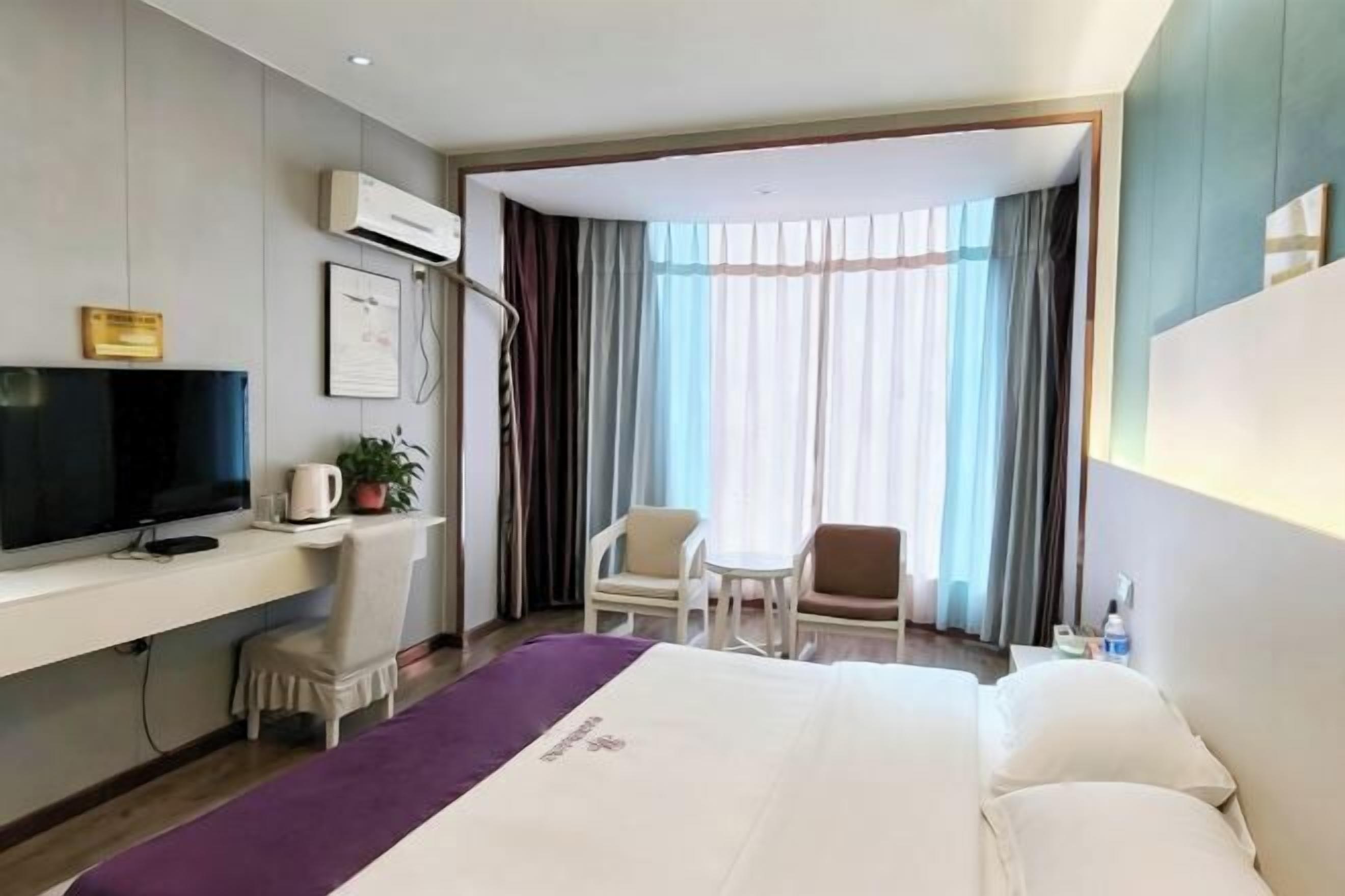 A hotel room with a bed, a desk with a TV, a chair, a small table, and a window with curtains.