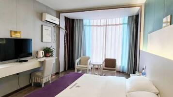 A hotel room with a bed, a desk with a TV, a chair, a small table, and a window with curtains.