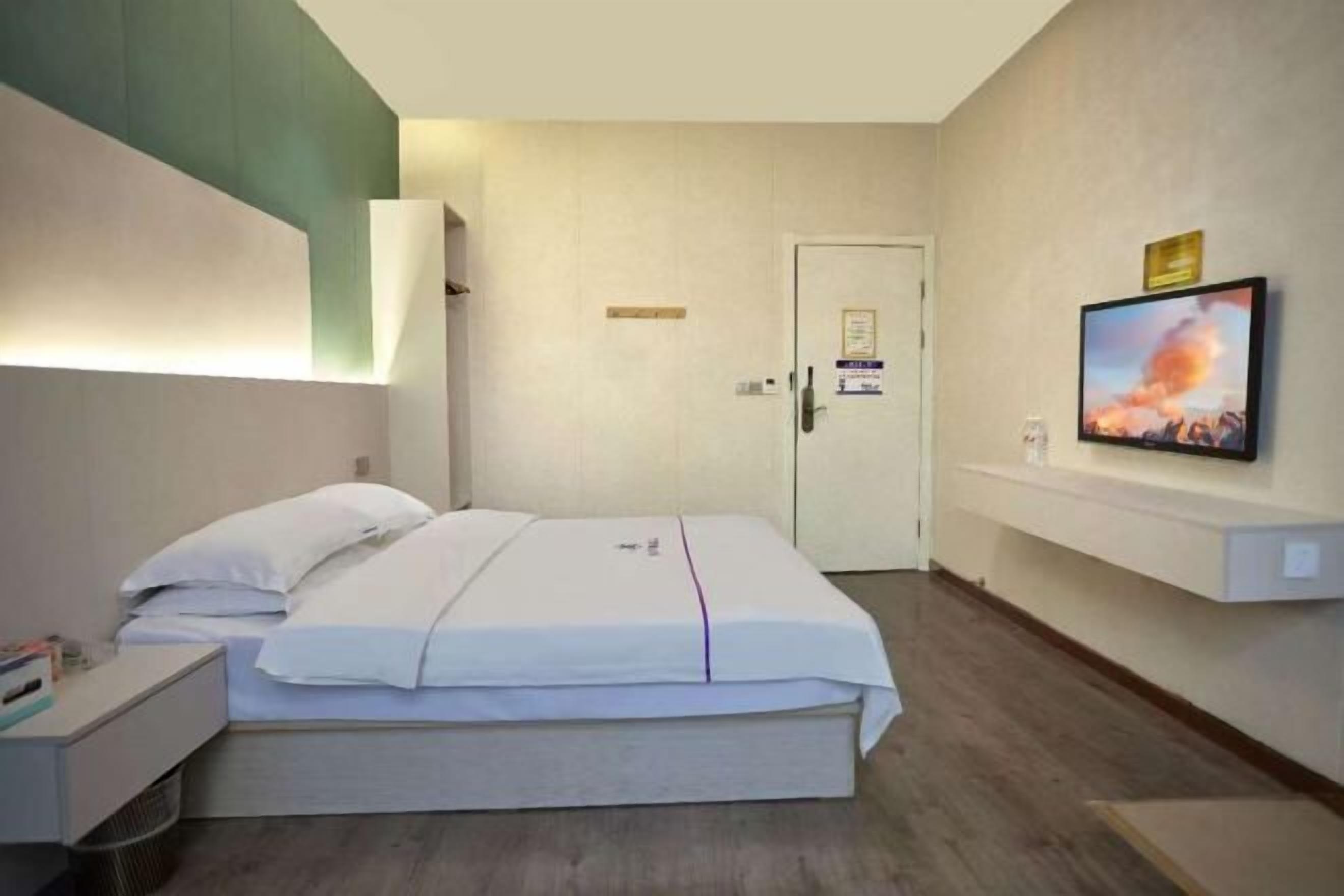 Hotellihuone, jossa on sänky, seinälle asennettu televisio, ovi ja pieni pöytä.