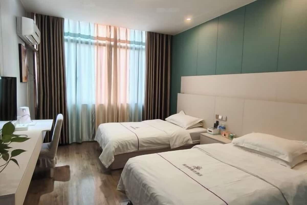 Una habitación de hotel con dos camas, un escritorio, una silla, un ventanal con cortinas y un equipo de aire acondicionado.