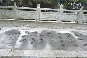 Una placa de piedra con texto grabado, una cerca de piedra con detalles decorativos y un camino pavimentado.