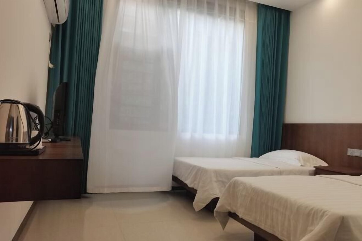 A hotel room with two beds, a wooden headboard, a desk with a lamp, and a window with sheer curtains.