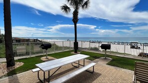 Un patio con una mesa de picnic blanca, dos parrillas y vista a la playa.