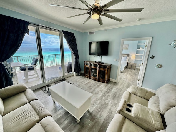 A spacious living room with a ceiling fan, a flat-screen TV, and a view of the ocean through large windows.