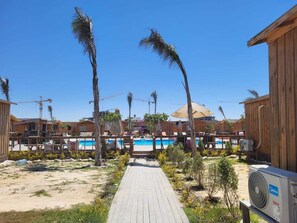 A wooden deck leading to a pool area with palm trees, a fan, and a clear blue sky.