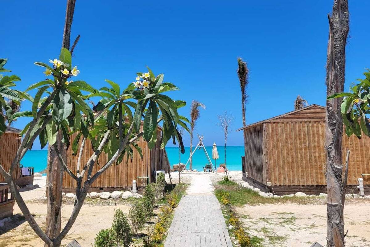 Een strandresort met houten hutten, een helderblauwe lucht en een zandpad dat naar het strand leidt.