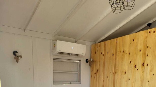 Une chambre avec un lit simple, un mur en bois, une petite fenêtre et un appareil de climatisation placé au-dessus du lit.