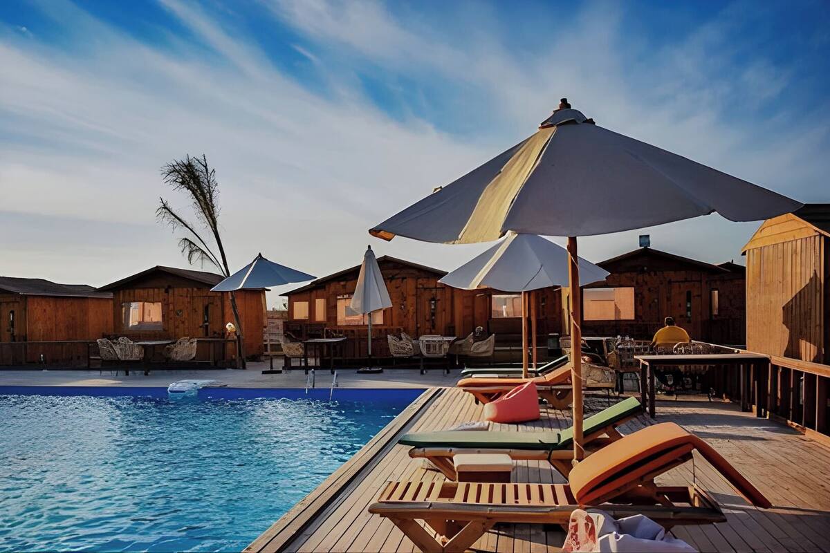 Een houten terras bij het zwembad met ligbedden en parasols, omgeven door houten huisjes.