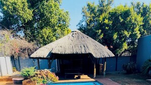 A rectangular swimming pool with clear blue water, surrounded by a brick patio and a thatched-roof structure.