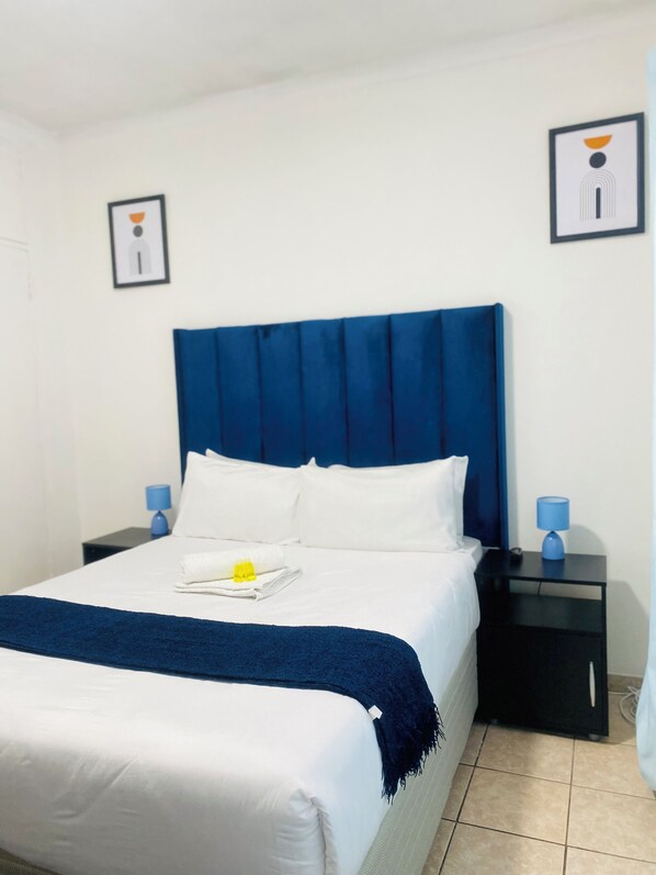 Un lit bien fait, avec une tête de lit bleue, du linge de lit blanc et un plaid bleu. Il y a deux tables de chevet avec des lampes, et la pièce présente un aménagement épuré et minimaliste.