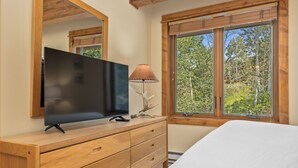 Une chambre à coucher avec une commode en bois, un téléviseur à écran plat, une lampe et une fenêtre donnant sur des arbres.