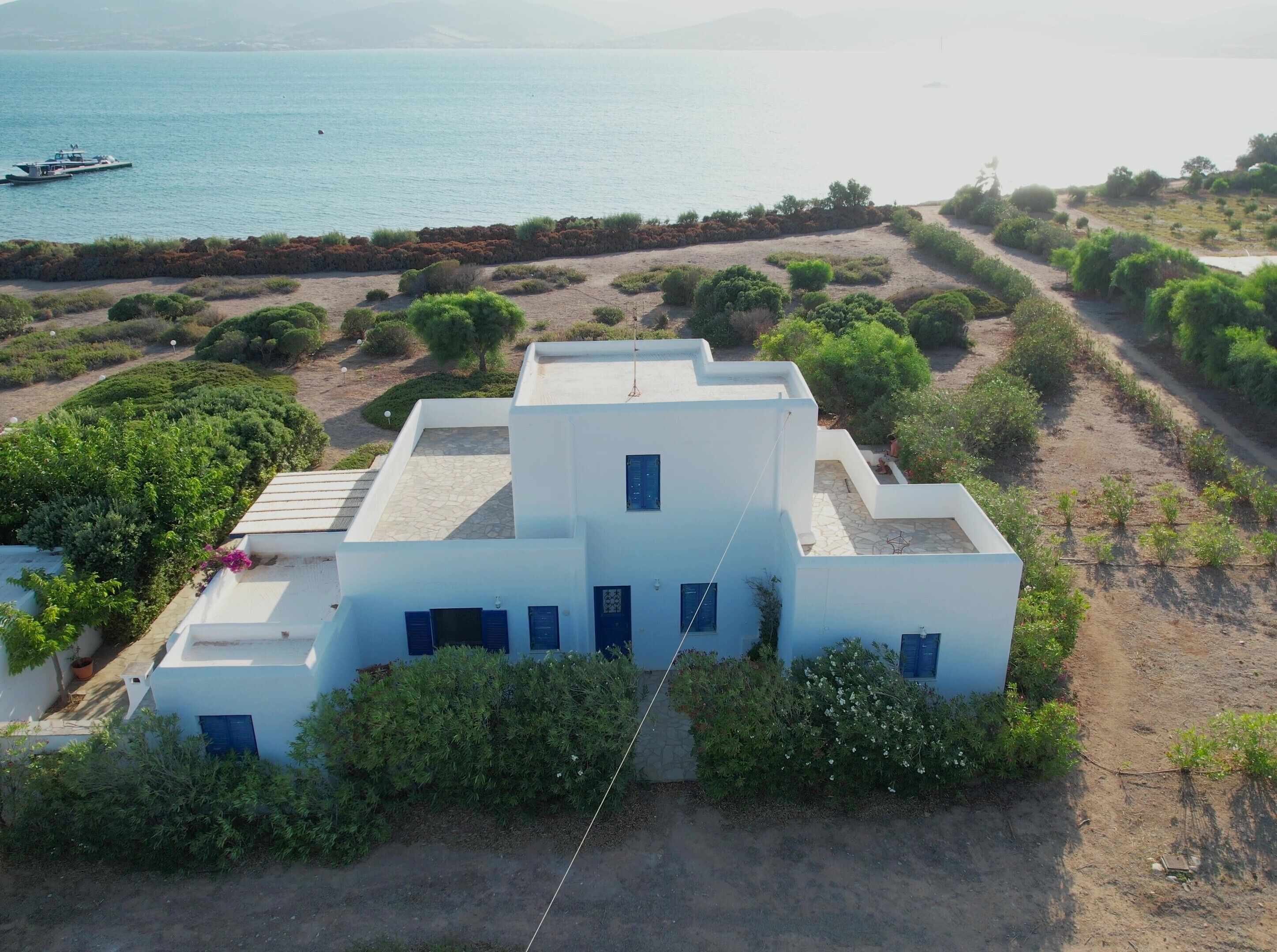 Un edificio blanco con techo plano y numerosas ventanas, rodeado de vegetación y un paisaje costero.