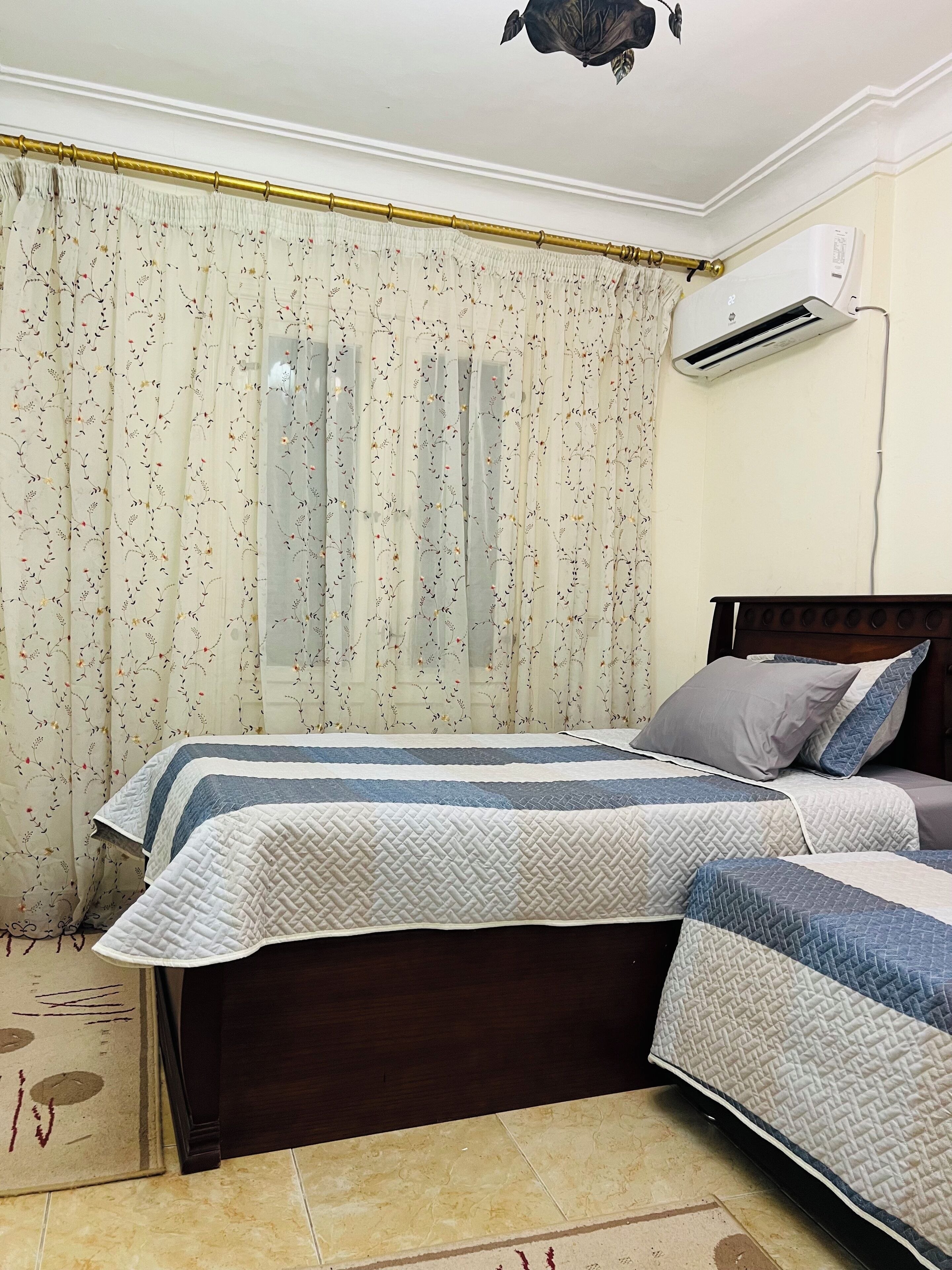 Un dormitorio con dos camas, cabecera de madera, aire acondicionado blanco y cortina con estampado floral.