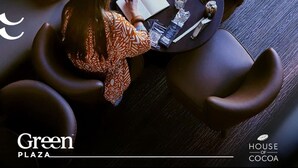 Eine Person sitzt an einem Tisch mit einem Notizbuch, einem Stift und einem Smartphone auf einer Ledersofa. Eine Handtasche liegt auf dem Sofa und ein Glas Wasser steht auf dem Tisch. Der Raum wirkt modern und bietet einen Ausblick durch ein Fenster.
