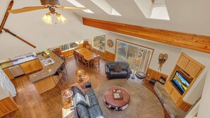 A spacious living area with a high ceiling, wooden beams, a dining table, a fireplace, and a television.