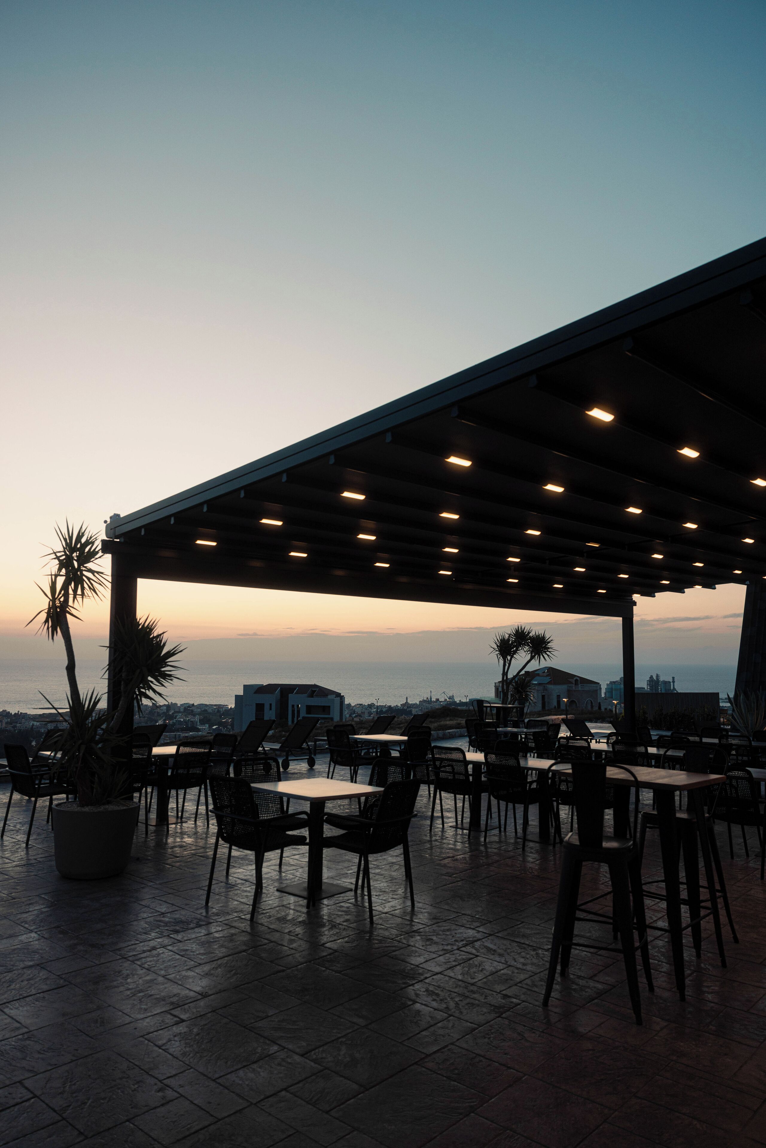 An outdoor patio area with tables and chairs, a pergola with lights, and a view of the ocean and cityscape at dusk.