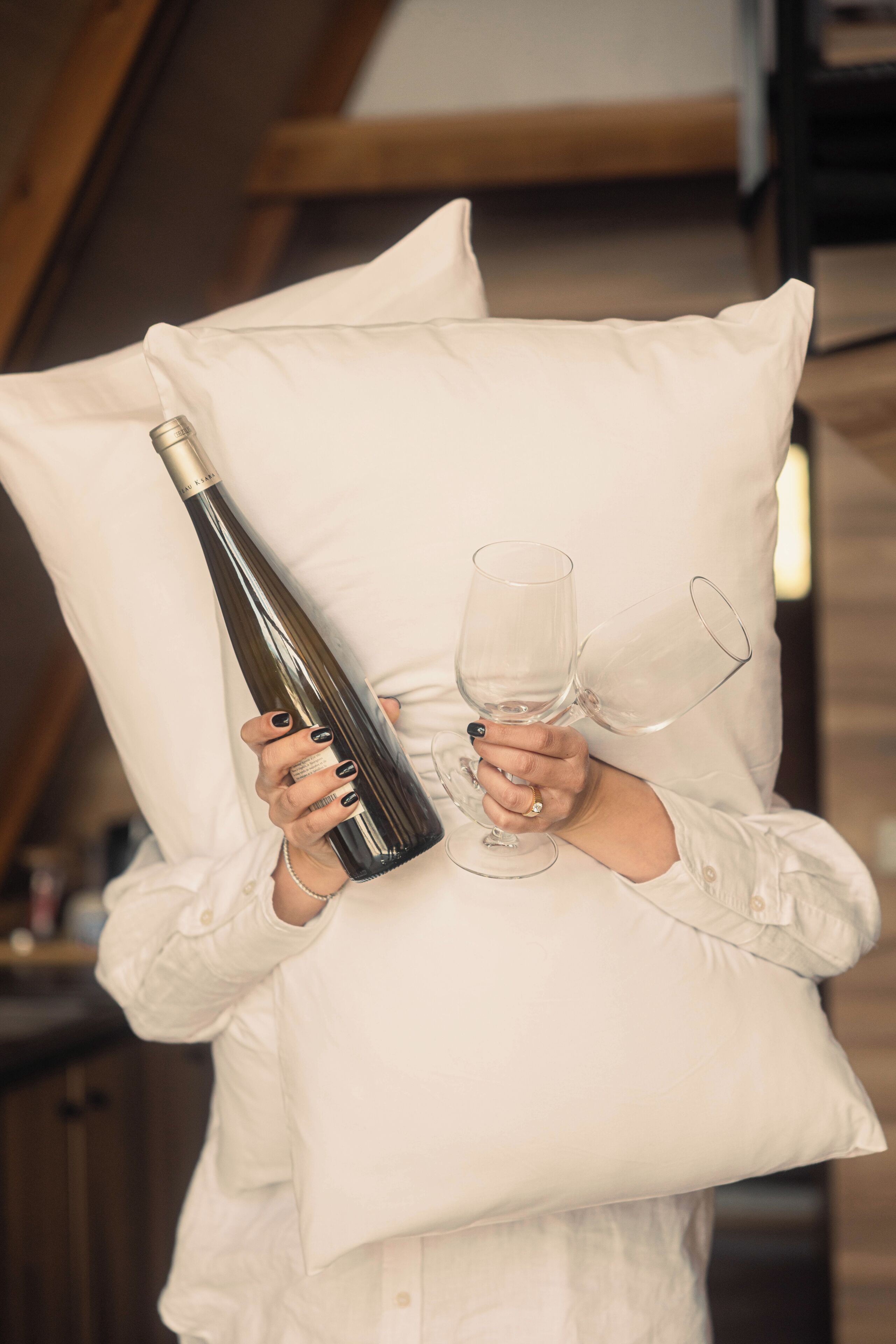 A person holding a wine bottle and two wine glasses, with a pillow in front of them.