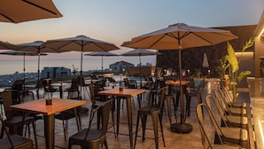 An outdoor dining area with multiple tables and chairs, umbrellas, and a pool in the background. - Panorama Lodges (Chekka)