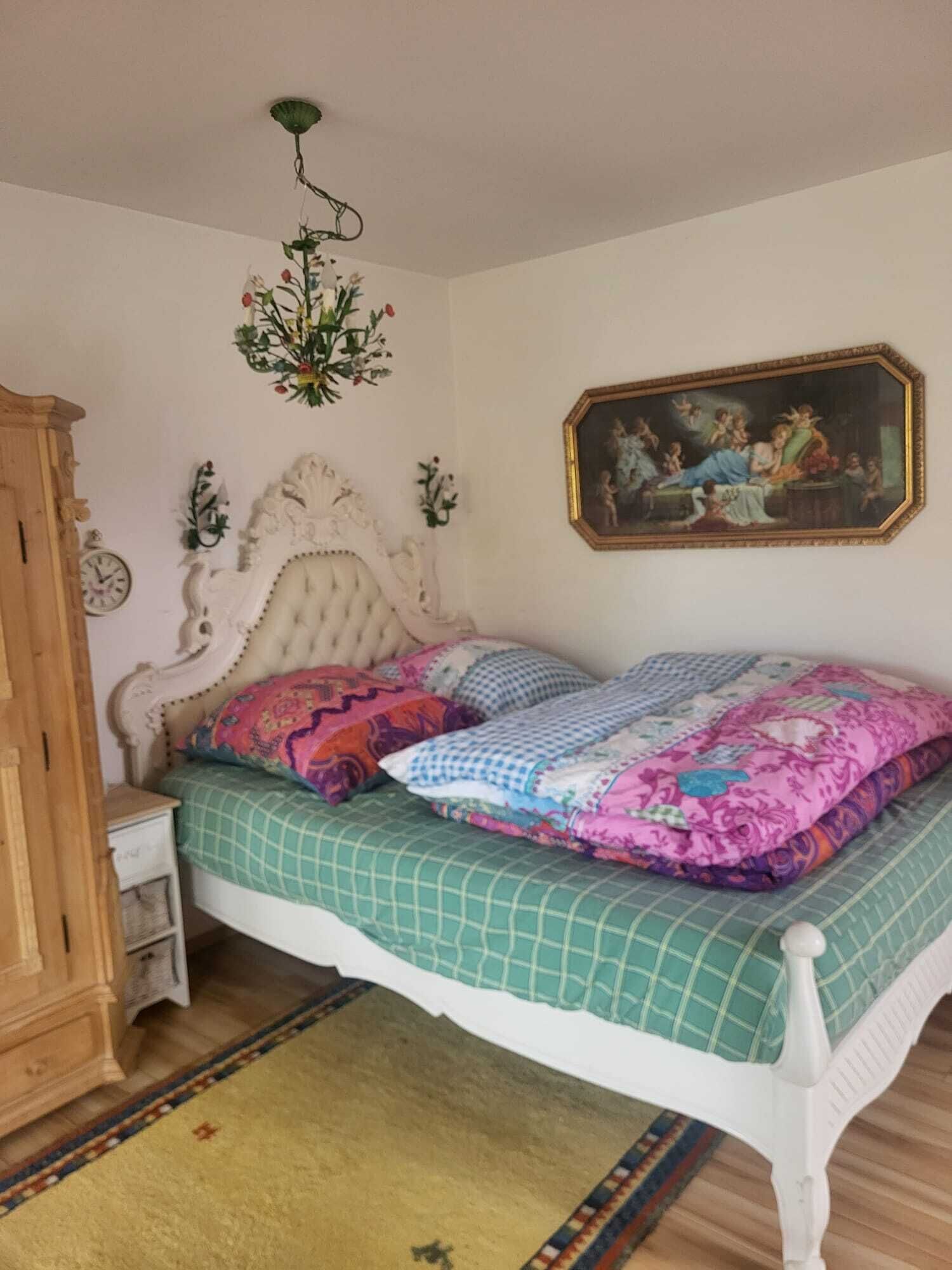 Une chambre à coucher avec un lit à baldaquin blanc, une couvre-lit à carreaux verts, un coussin à motifs floraux, un tableau accroché au mur et une table de chevet en bois.