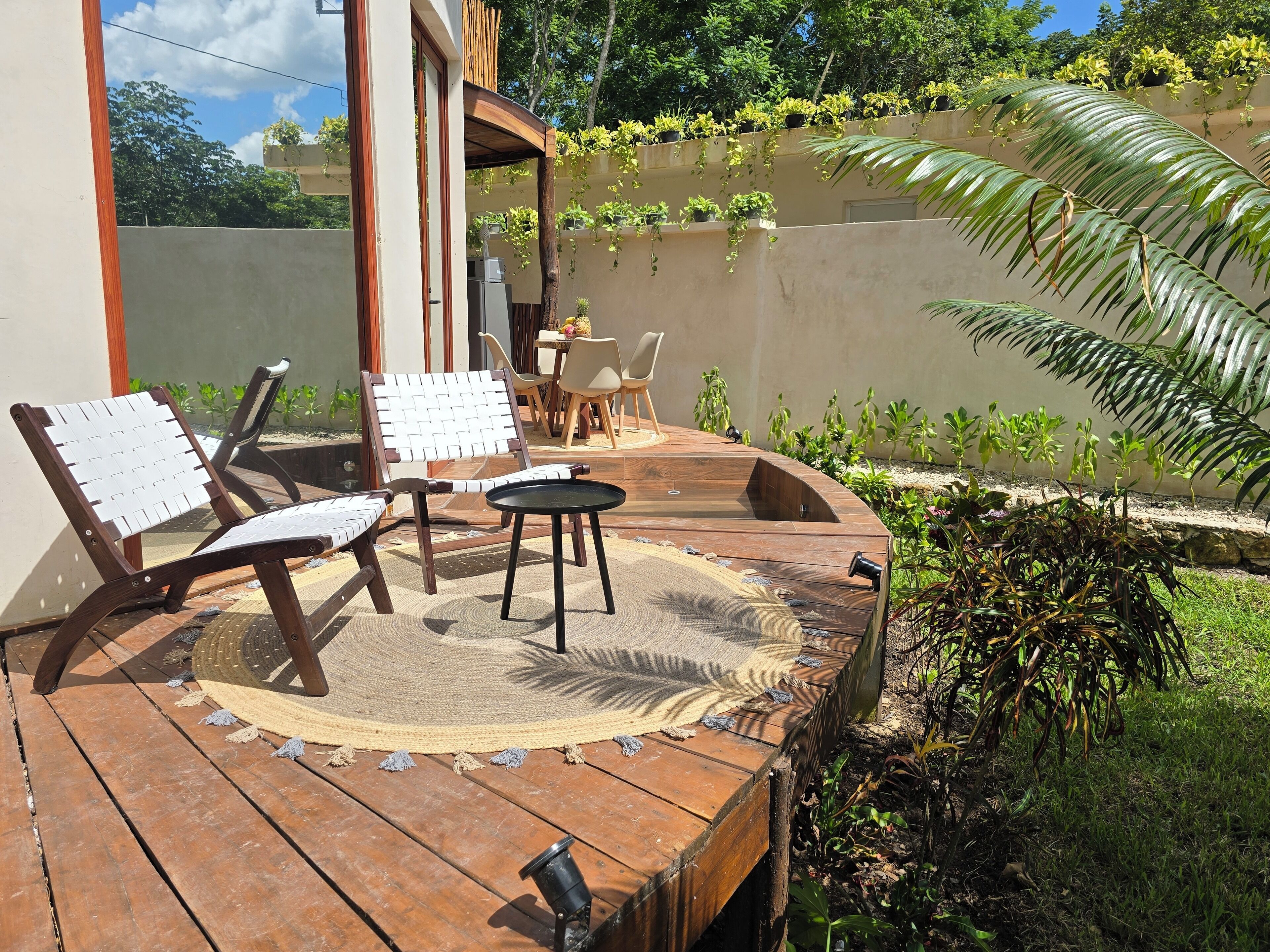 Una terraza de madera con dos sillas y una mesita, rodeada de vegetación y un edificio con barandilla de madera.