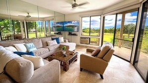 A spacious living room with a large sofa, armchairs, and a coffee table. There are two flat-screen TVs mounted on the wall, and a ceiling fan. The room has large windows offering a view of a green landscape.