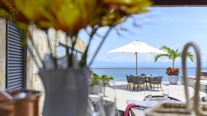 Ein Essbereich im Freien mit Tischen und Stühlen, einem weißen Sonnenschirm und Blick auf das Meer.