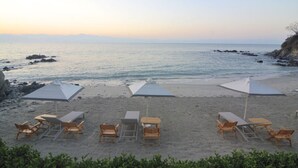 Ein Strand mit Liegestühlen und Sonnenschirmen, freie Sicht auf das Meer und Felsformationen.