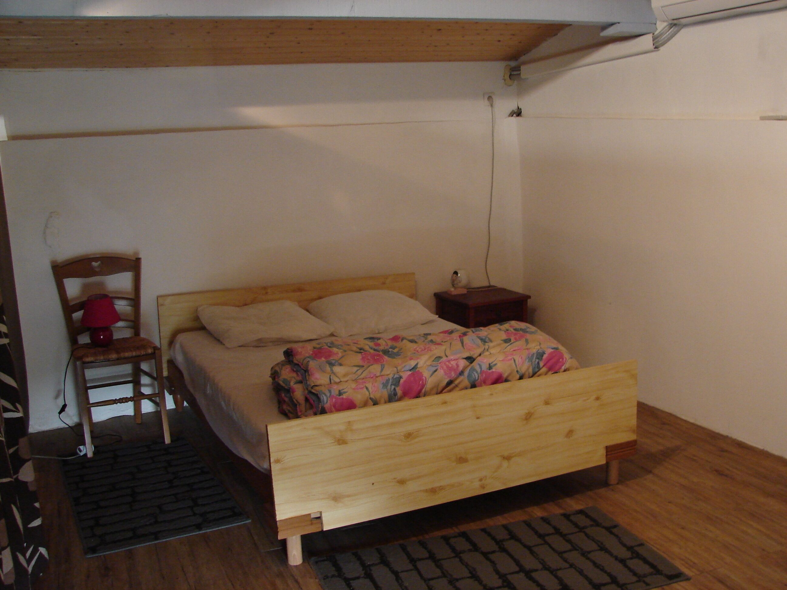 Una cama individual con estructura de madera y colcha estampada, una mesita de noche de madera con una lámpara, y una silla con lámpara roja.