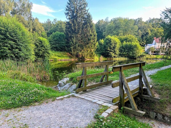 A wooden bridge over a pond in a park with dense trees and a building in the background. - Famielienapartment "An der Forellenschänke " (Sebnitz)