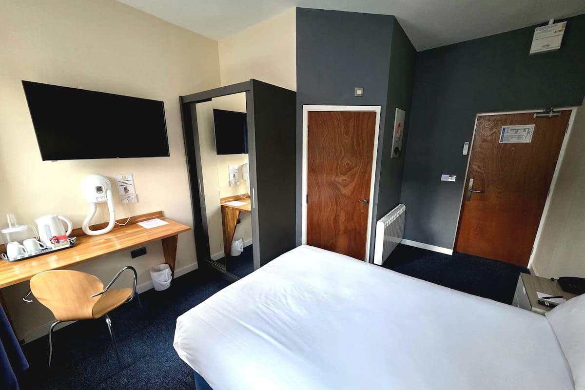 A hotel room with a bed, a desk with a coffee maker, a chair, a mirror, and a wardrobe.