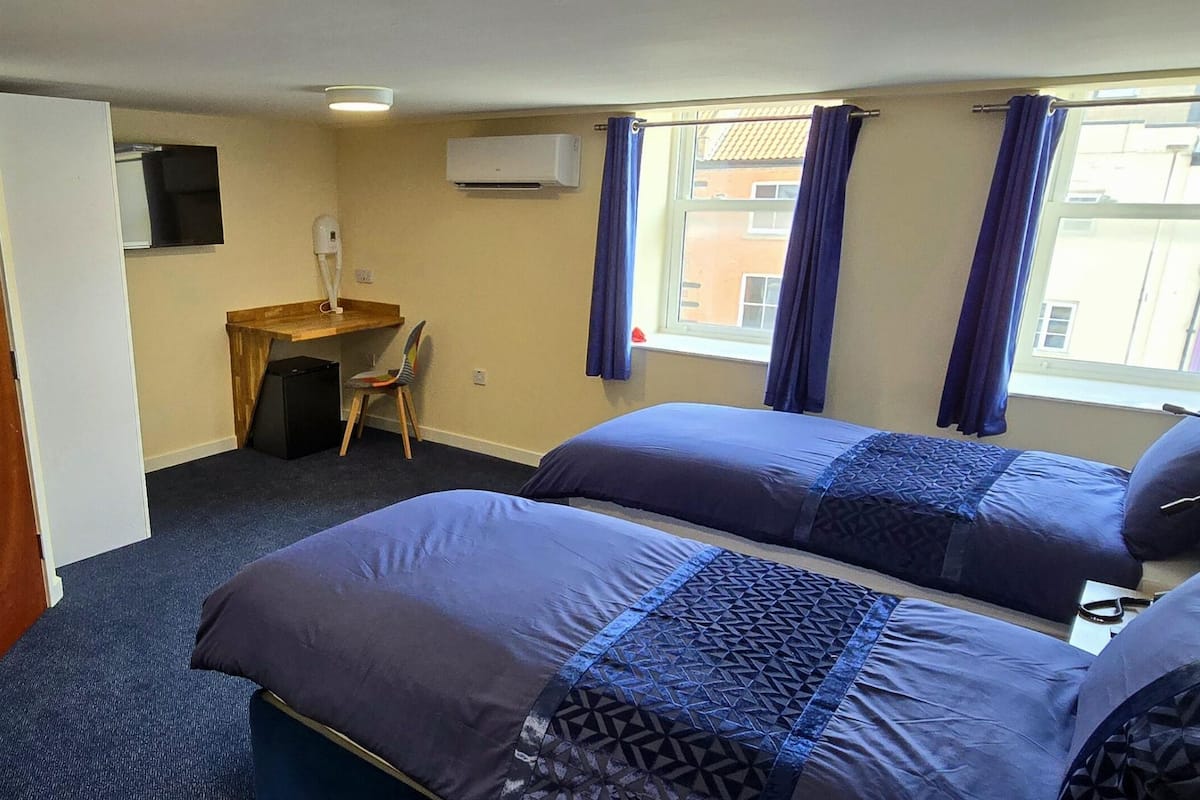 A hotel room with two beds, a desk, a chair, an air conditioning unit, and a window with curtains.