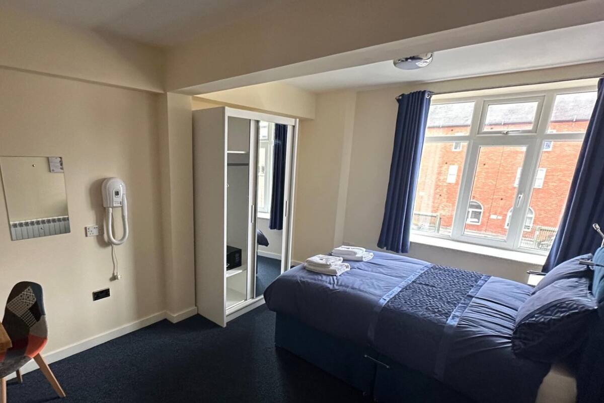 A hotel room with a bed, a desk with a chair, a wardrobe, a window with curtains, and a telephone on the wall.