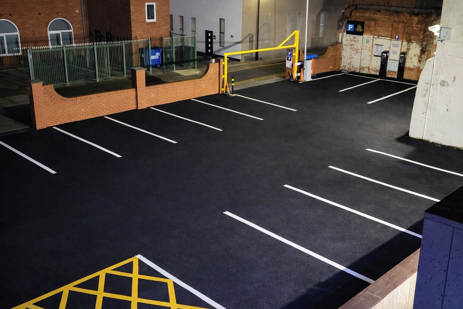 Un parking avec des places délimitées, une barrière de sécurité jaune et un bâtiment en briques en arrière-plan.
