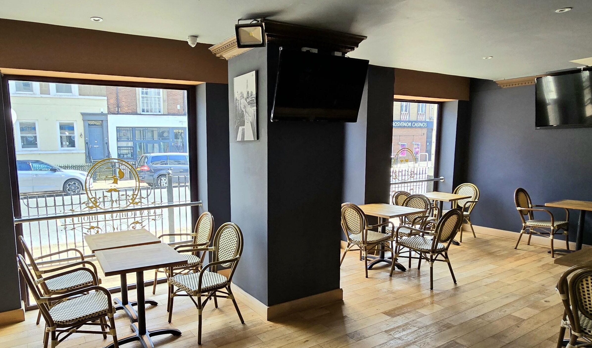 An indoor seating area with wicker chairs and tables, large windows with a view of the street, and a mounted television on the wall.