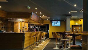 A bar area with a counter, stools, and a television screen displaying a scene. - The Carmichael - Hull City Centre (Hull)