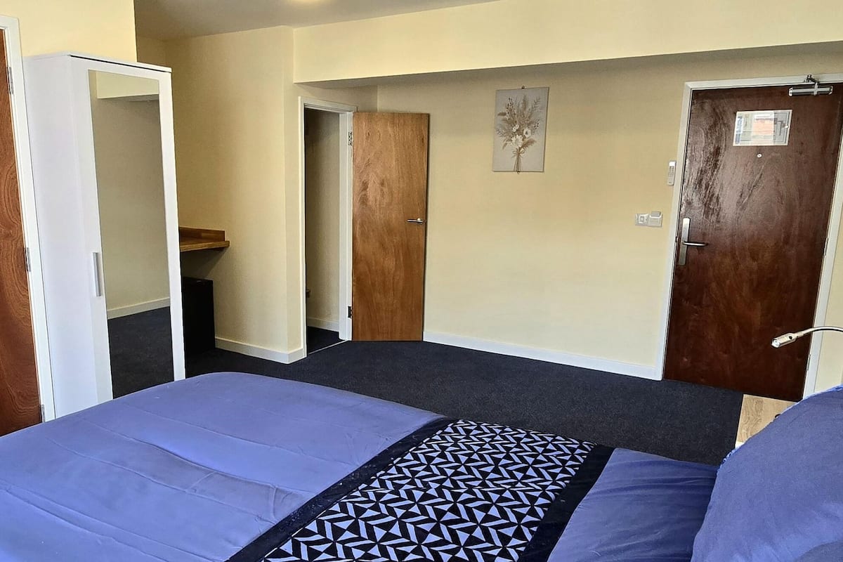 A hotel room with a bed, a wooden door, a small bathroom, and a hallway leading to another room.