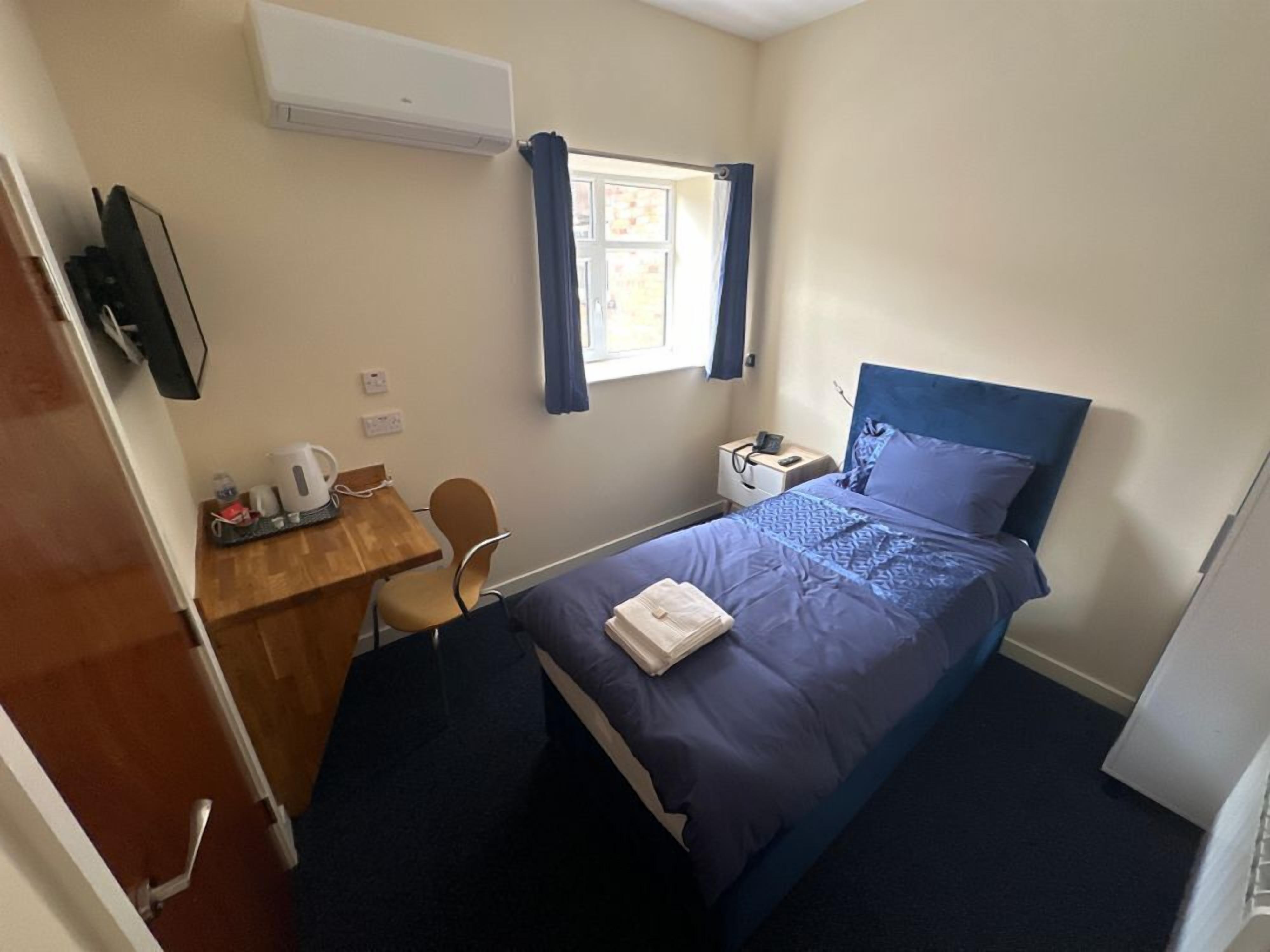 A small, neatly arranged bedroom with a single bed, a desk with a chair, a window with curtains, and a wall-mounted air conditioner.