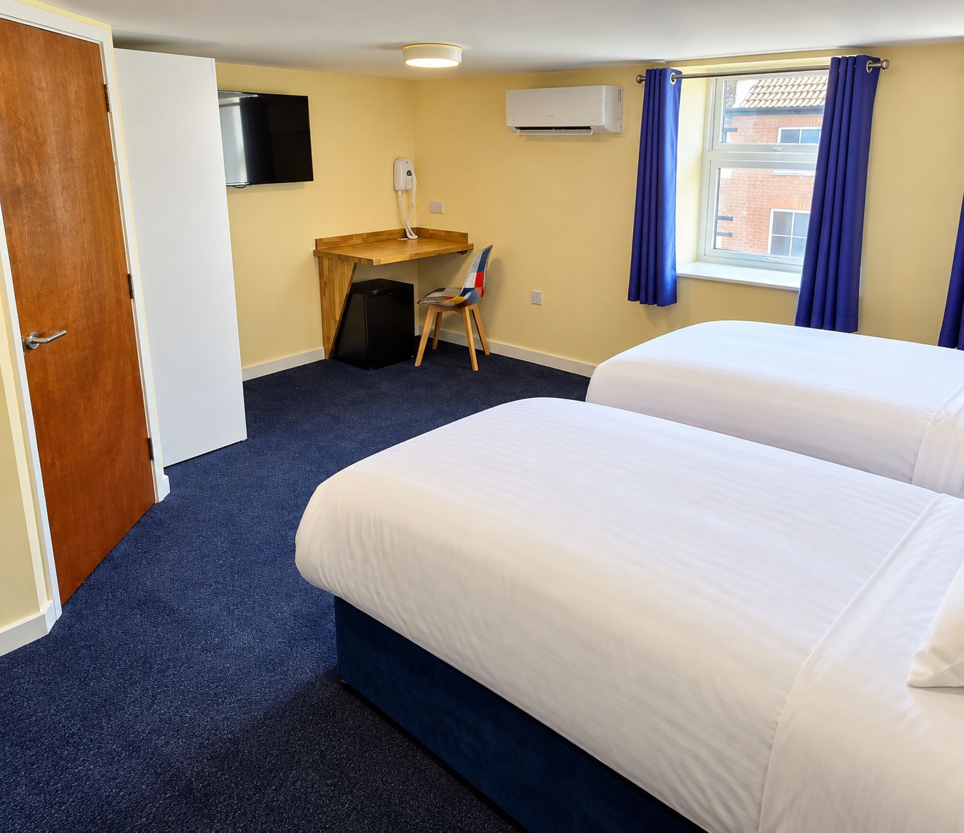 A hotel room with two beds, a desk, a chair, a flat-screen TV, and a window with curtains.