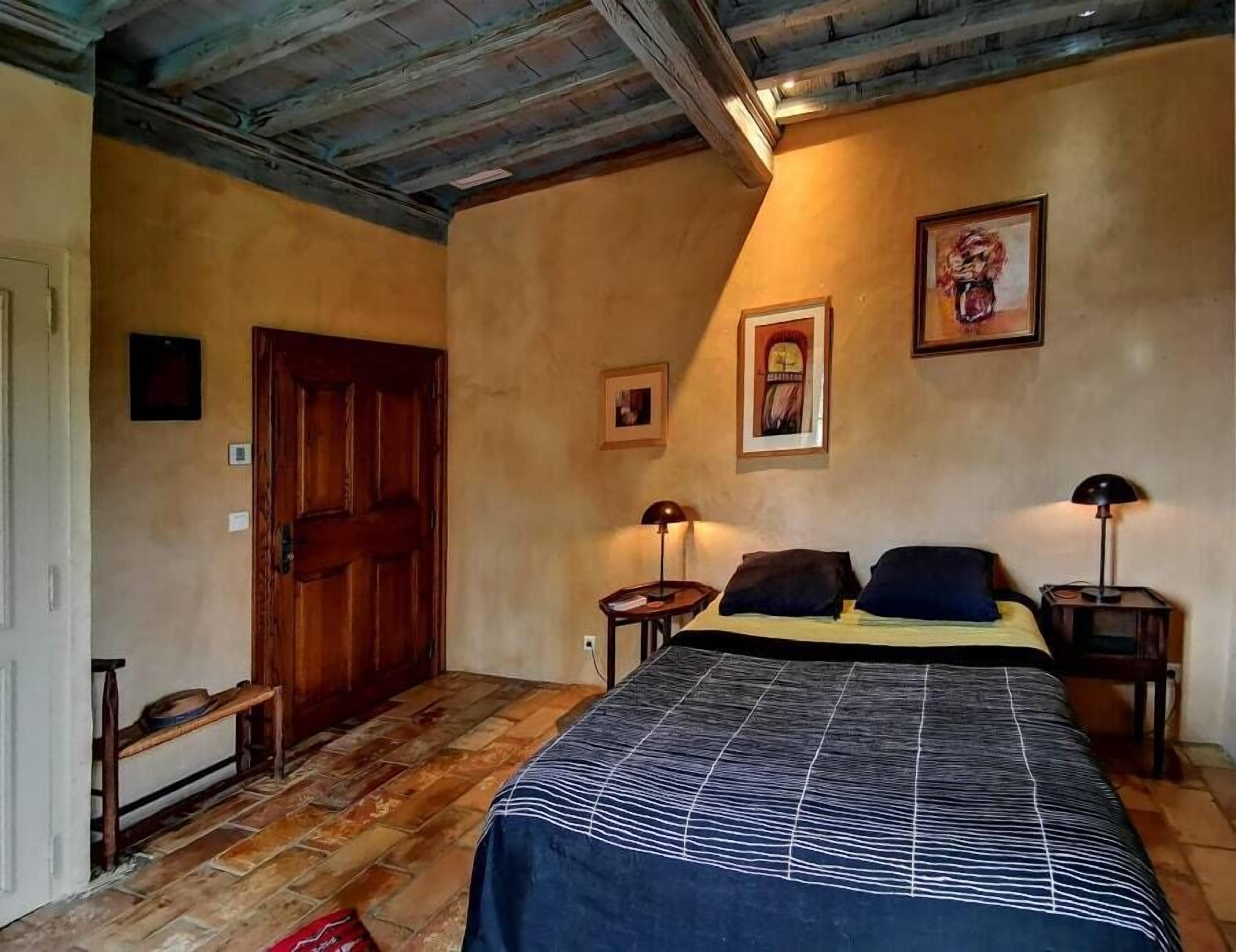 Une chambre avec une porte en bois, un lit recouvert d’une couvre-lit à motifs, deux tables de chevet avec des lampes et un tableau encadré au mur.
