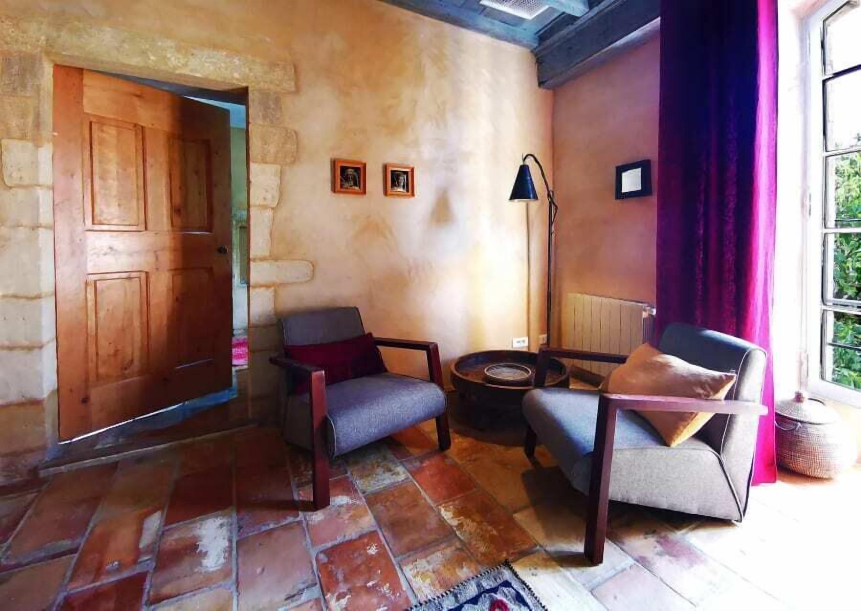 A cozy room with a stone wall, a wooden door, two armchairs, a small round table, and a floor lamp.