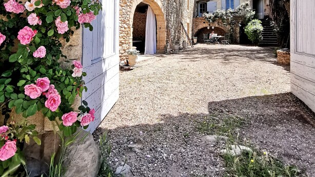 A stone building with an arched entrance, a gravel courtyard, and pink flowers in the foreground. - La Bastide du Farfadet (Laudun)
