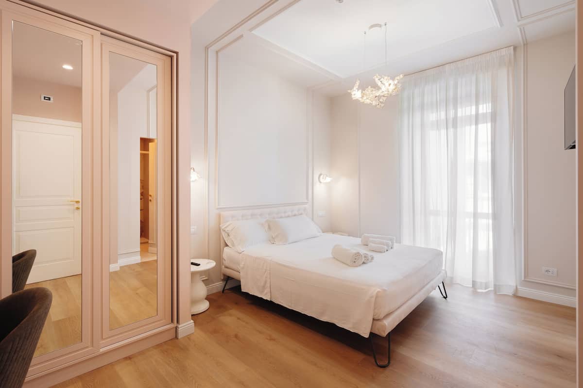 Een slaapkamer met een grote spiegel, een bed met witte lakens, een nachtkastje en een kroonluchter.