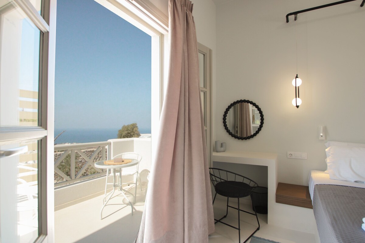 A balcony with a view of the sea, a small table, a chair, and a bed with white bedding.