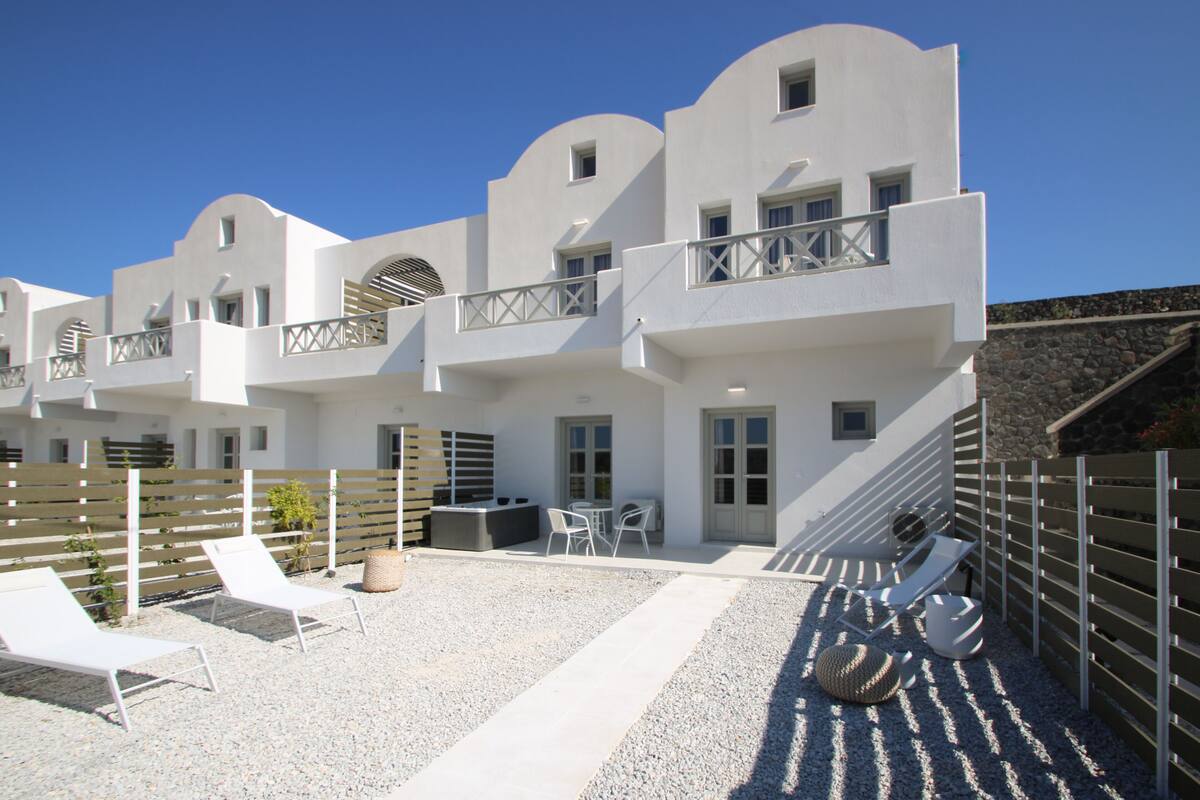 Un bâtiment moderne de couleur blanche, doté de nombreux balcons et d’une terrasse en gravier aménagée avec des chaises longues et une table.
