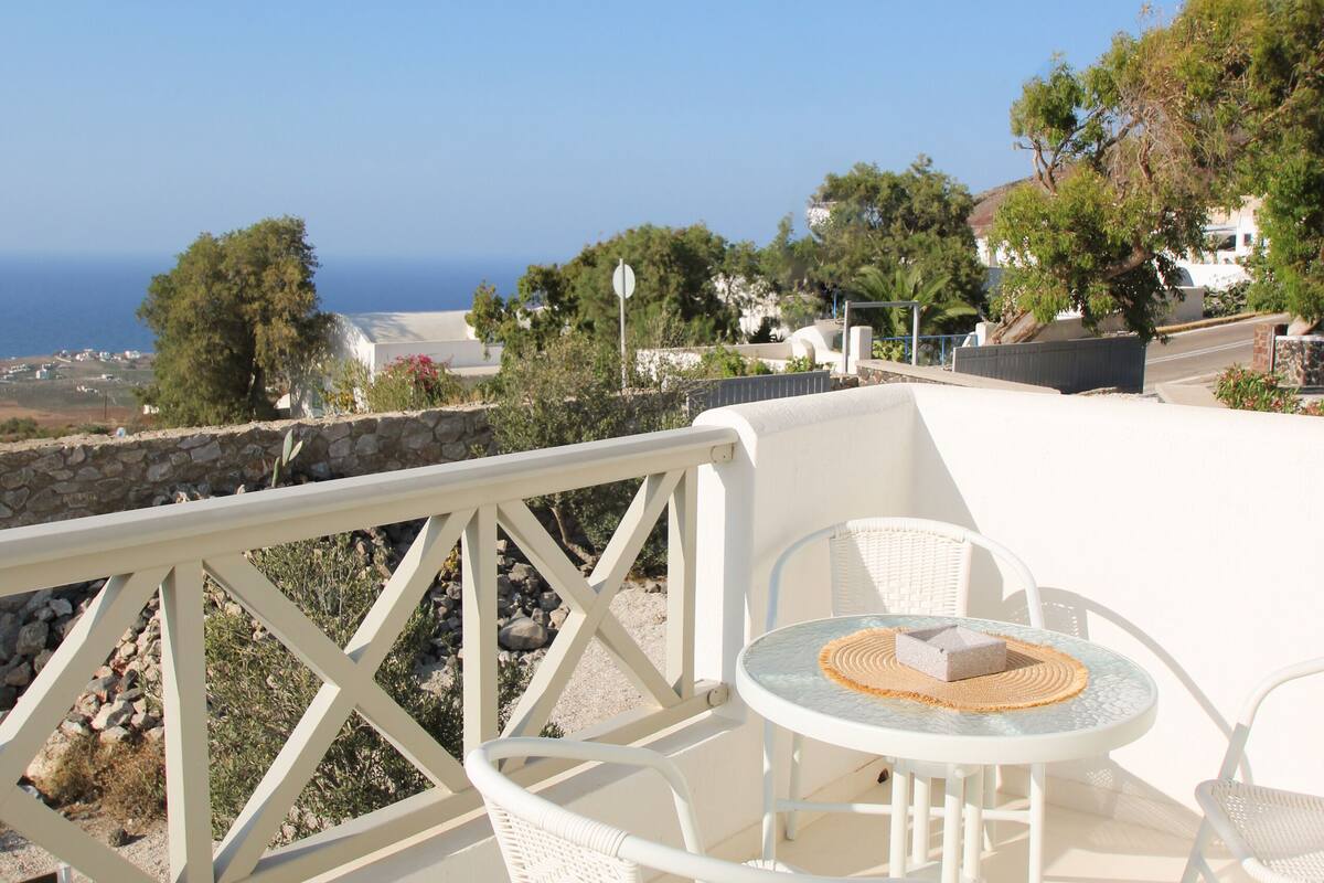 Une terrasse dotée de meubles en osier blanc et d’une table, offrant une vue sur un paysage côtier avec des maisons et des arbres.