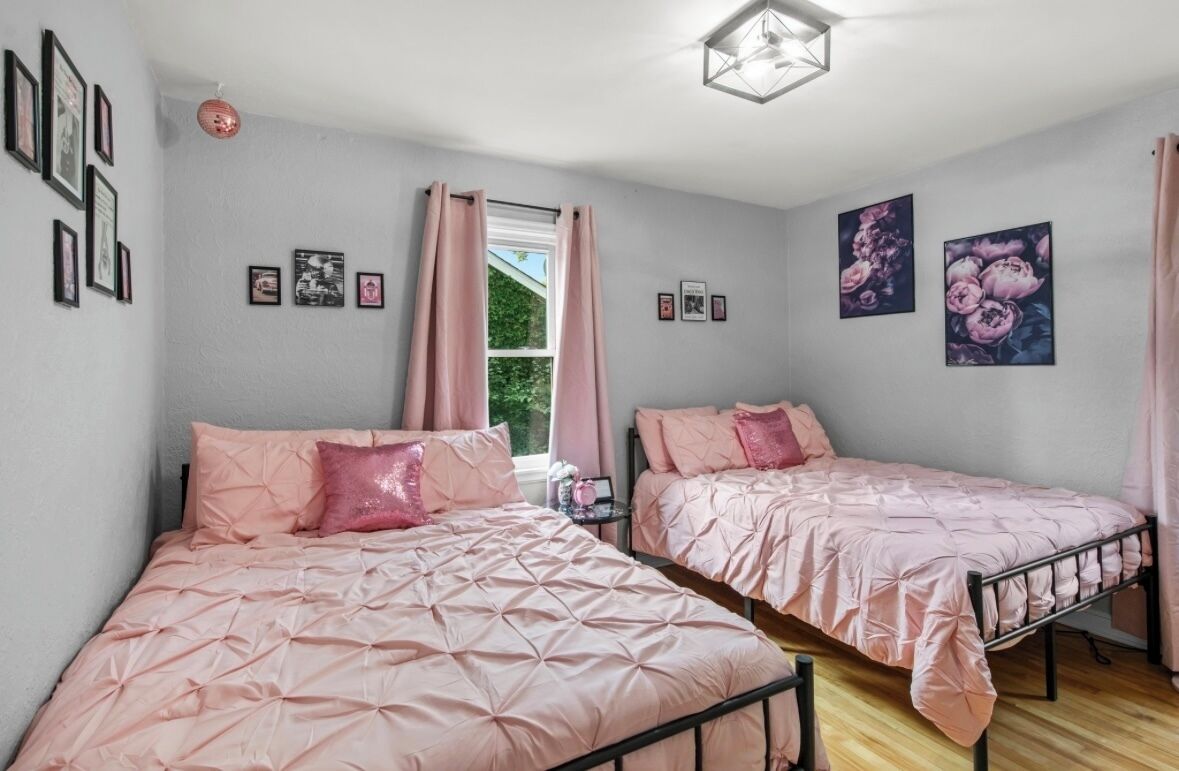 Ein Schlafzimmer mit zwei Betten, jeweils mit rosa Bettwäsche und Kissen, einem Fenster mit Vorhängen und Wandbildern.