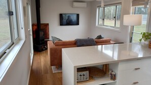 A modern living room with a sofa, a fireplace, a TV, and a kitchen area with a toaster. - Lakeside Off-Grid Luxury Cabin Redman View (Lake Fyans)