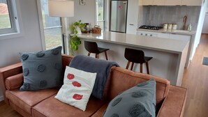 A modern living room with a brown leather sofa, a small kitchen area with a gas stove, and a dining table with chairs. - Lakeside Off-Grid Luxury Cabin Redman View (Lake Fyans)